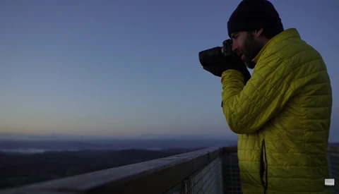 Photographer in yellow jacket shooting at dusk from elevated vantage point overlooking landscape.