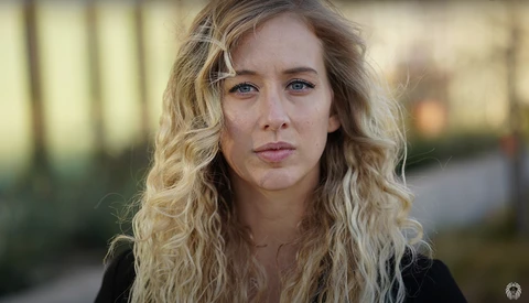 Woman with wavy blonde hair looking directly at camera with neutral expression outdoors.