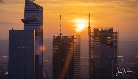 Golden sunrise illuminates modern skyscrapers with construction cranes silhouetted against the sun.