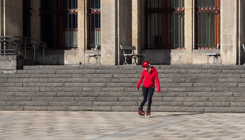 Person in red jacket rollerskating across a wide stone plaza in front of a modernist building.