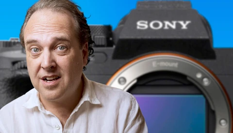 Man in white shirt gesturing expressively toward Sony mirrorless camera against blue background.