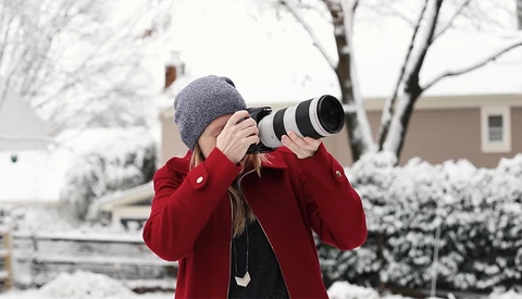 Child in red coat and gray beanie photographing with telephoto lens in snowy residential setting.