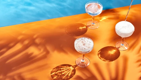 Cocktails suspended above an orange fabric with blue water and palm leaf shadows in bright sunlight.