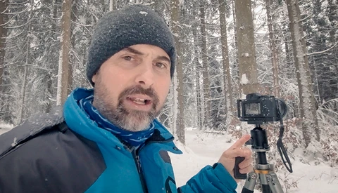Videographer holding a gimbal stabilizer while filming in a snowy forest setting.