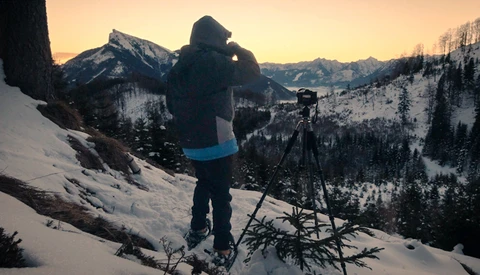 Photographer with camera on tripod overlooking snow-covered mountain landscape at golden hour.