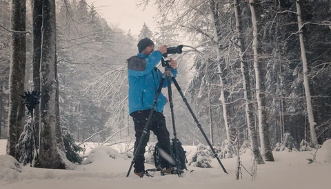 Photographer in blue jacket shooting with telephoto lens on tripod in snowy forest.