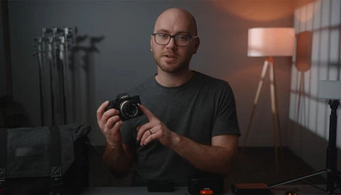 Photographer holding a mirrorless camera in a studio setting with warm window lighting.