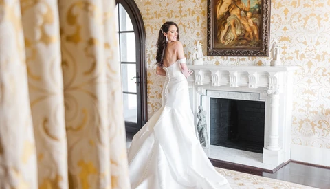 Bride in off-shoulder wedding dress with train standing by ornate fireplace in classical interior.