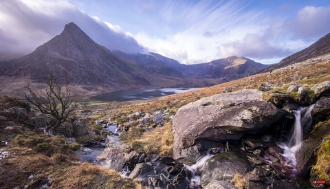 Mountain valley landscape with cascading waterfall, alpine lake, and dramatic peaked peaks under cloudy sky.