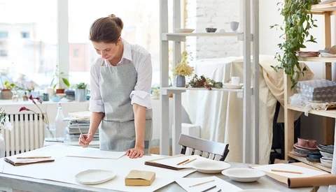 Person leaning over a desk arranging ceramic plates and printed materials in a bright studio space.