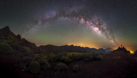 Milky Way galaxy arcing across night sky above desert landscape with mountains and distant lights.