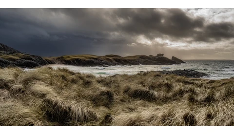 Windswept coastal dunes with marram grass overlooking a dramatic seascape under stormy skies.