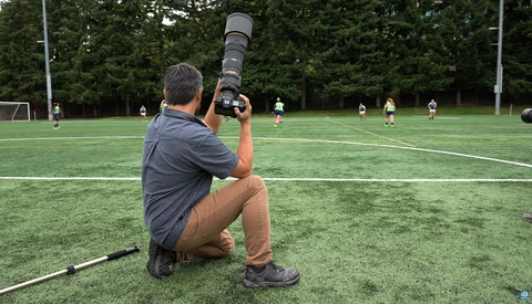 Sports photographer crouching on artificial turf with telephoto lens aimed at youth soccer match.
