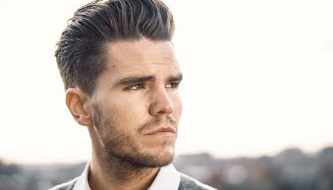 Male portrait with styled pompadour hair and beard, looking away from camera against blurred outdoor background.