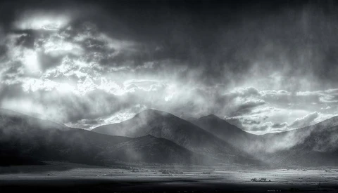 Dramatic black and white landscape with a solitary mountain rising from a vast plain beneath stormy clouds.