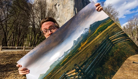 Photographer holding a printed landscape photograph outdoors, comparing the print to the actual mountain valley scene behind it.
