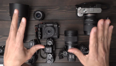 Flat lay of photography equipment including cameras, lenses, and accessories arranged on dark wood with hands framing the composition.