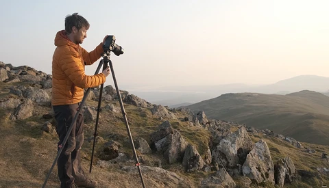 Photographer on rocky hilltop operating camera mounted on tripod during golden hour.