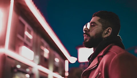 Man wearing glasses and maroon jacket against neon pink signage and teal night sky.
