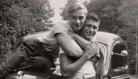 Two men posing playfully on the hood of a vintage automobile in a wooded setting.