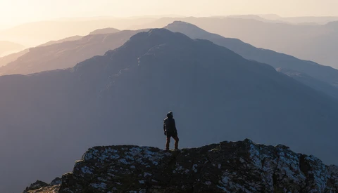 Silhouetted hiker standing on a rocky outcrop with layered mountains in atmospheric haze beyond.