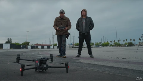 Two people stand on an empty parking lot with a drone in the foreground, operating remote controls under overcast skies.