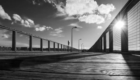 Empty concrete pathway between tall mesh fencing under bright sunlight and dramatic clouds.