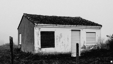 Deteriorating white concrete structure with dark shuttered openings and moss-covered roof in rural landscape.
