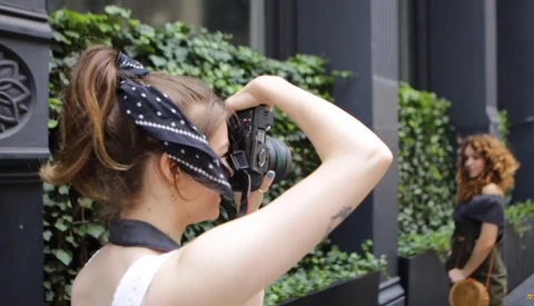 Photographer holding a camera to her face while shooting, with a model visible in the background near ivy-covered wall.