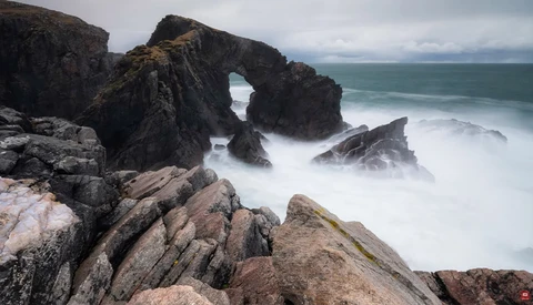 Rocky coastal outcrop with natural stone arch framing turbulent ocean swells and misty waves.