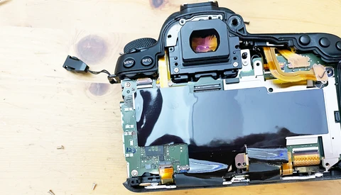 Disassembled camera showing internal circuitry, mirror mechanism, and sensor components on wooden surface.