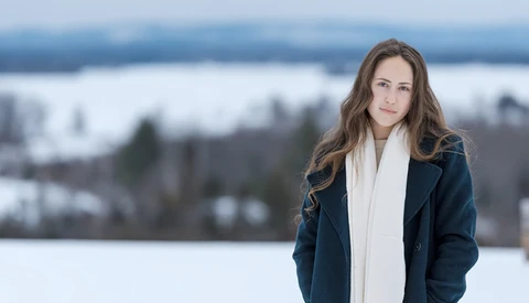 Woman in dark jacket and white scarf standing outdoors with snowy landscape behind her.