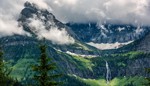 Alpine valley with forested slopes, a cascading waterfall, and dramatic peaks shrouded in low clouds.