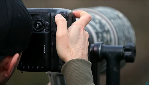 Photographer holding a Canon mirrorless camera in shooting position, with telephoto lens visible in background.
