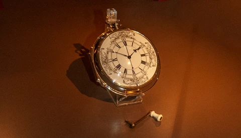 Ornate pocket watch with Roman numerals and zodiac symbols on cream dial, photographed against warm brown background with dramatic side lighting.