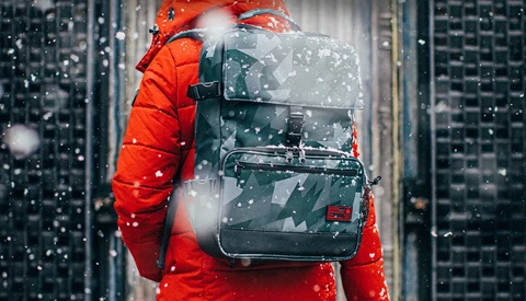 Person wearing red jacket and backpack standing in urban setting with falling snow.