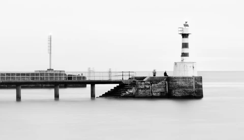 Lighthouse at the end of a long pier extending into calm water, captured in black and white.