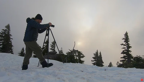 Photographer using a tripod-mounted telescope on snow-covered ground with coniferous trees and overcast sky.