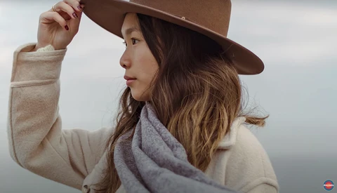 Woman in cream sweater and brown hat adjusting her hat brim while looking to the side.