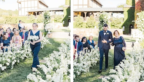 Wedding ceremony with guests walking through white flowering plants lining an outdoor garden aisle.