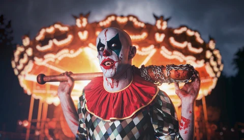 Clown performer in full theatrical makeup holding a baseball bat against an illuminated carnival ride backdrop.