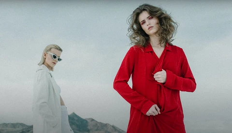 Two women standing against a mountainous landscape under overcast sky, one wearing a red jacket and the other in neutral tones with sunglasses.