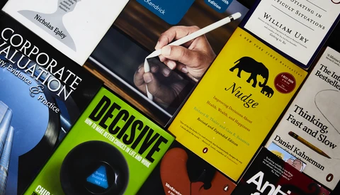 Hand holding a stylus over an open business book surrounded by stacked professional development books on a dark surface.