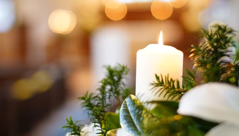 Lit white pillar candle surrounded by green foliage and white flowers with warm bokeh lights in soft focus background.
