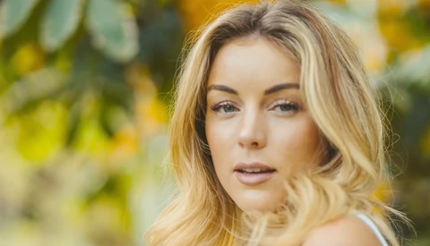 Woman with blonde wavy hair looking directly at camera with soft, natural lighting and blurred green foliage background.