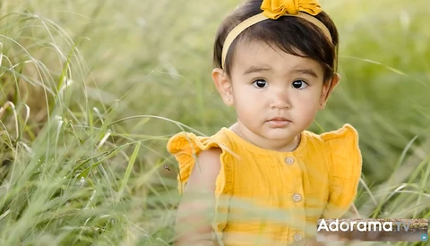 Toddler in yellow dress with matching headband sitting in tall grass during outdoor portrait session.