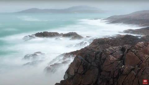 Rocky coastline with turbulent waves and misty long-exposure effect under overcast sky.