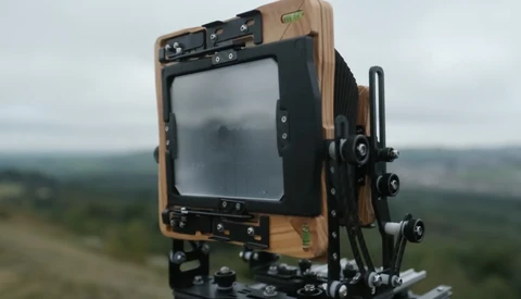 Large format camera with wooden body and bellows mounted on tripod, photographing a misty forested landscape.