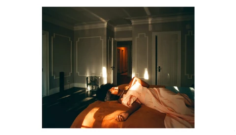 Sunlit bedroom with unmade bed featuring orange bedding and white walls with paneling.