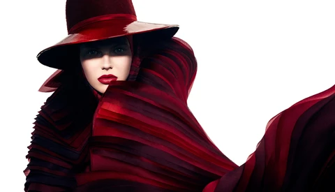 Portrait of a person wearing a dramatic red and burgundy draped garment and wide-brimmed hat against white background.